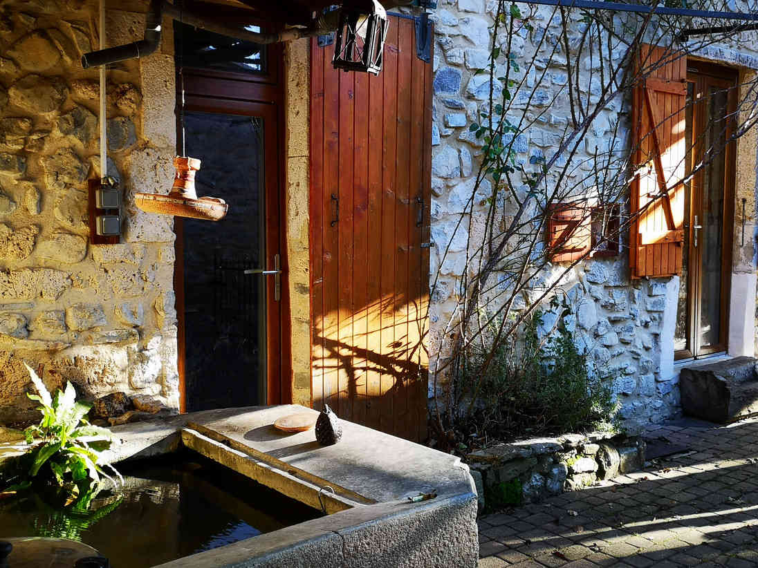 Une cour ensoleillée avec une porte en bois, des fenêtres à volets en bois, un mur en pierre et une fontaine en pierre