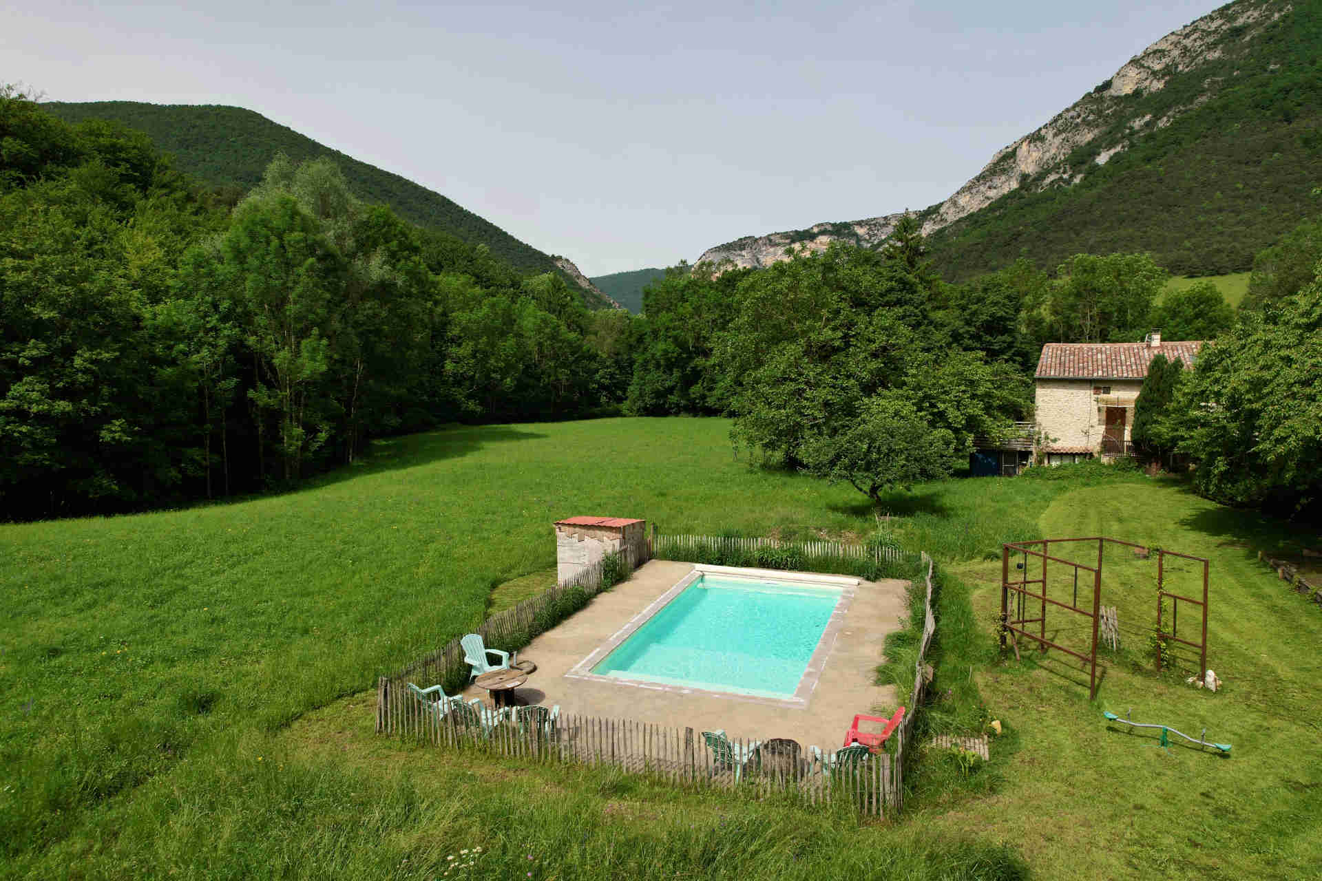 Piscine rectangulaire entourée d’une clôture en bois au premier plan. L’arrière-plan présente des champs et des arbres verdoyants, avec des montagnes qui s’élèvent au loin sous un ciel clair.