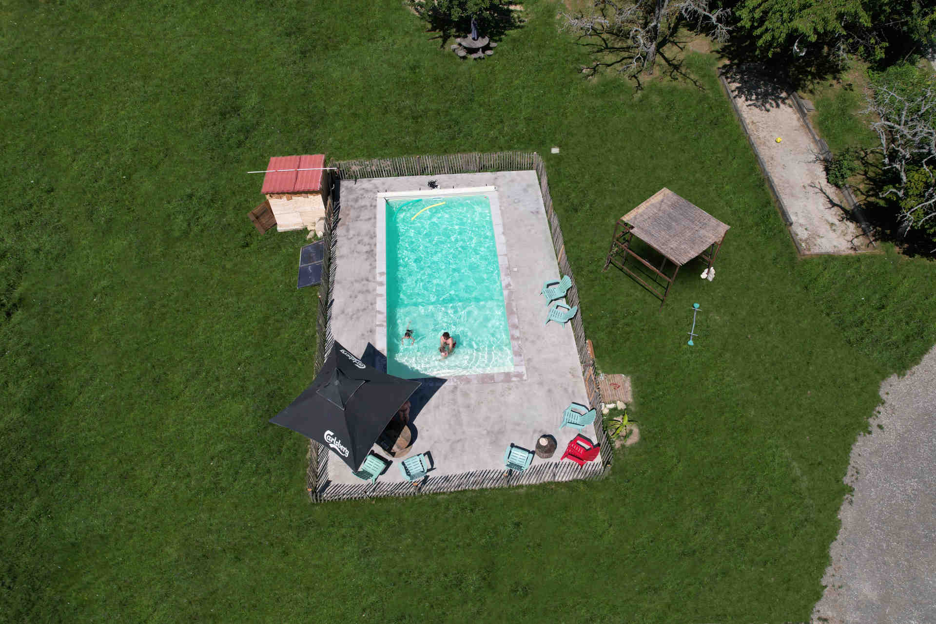 Vue aérienne d'une piscine rectangulaire entourée de chaises longues, un parasol, et de l'herbe verte, avec deux personnes se baignant.