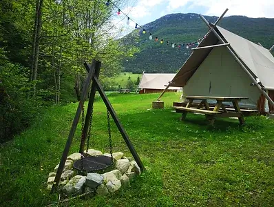 Un campement pittoresque avec une tente de style tipi, un foyer, et une table de pique-nique, situé dans une vallée verdoyante avec des montagnes majestueuses en arrière-plan. L'ambiance est tranquille et en harmonie avec la nature.