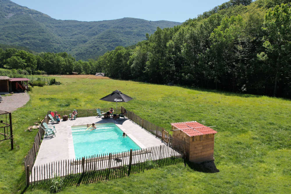 Une piscine extérieure rectangulaire entourée d’une clôture en bois, avec des personnes se détendant sur des chaises longues et dans l’eau. Un parasol noir avec un logo est à côté de la piscine. La piscine est située dans un espace vert ouvert avec une pelouse bien entretenue et une chaîne de montagnes couverte d’arbres verts luxuriants en arrière-plan. Une petite structure avec un toit rouge est visible à droite de la piscine.