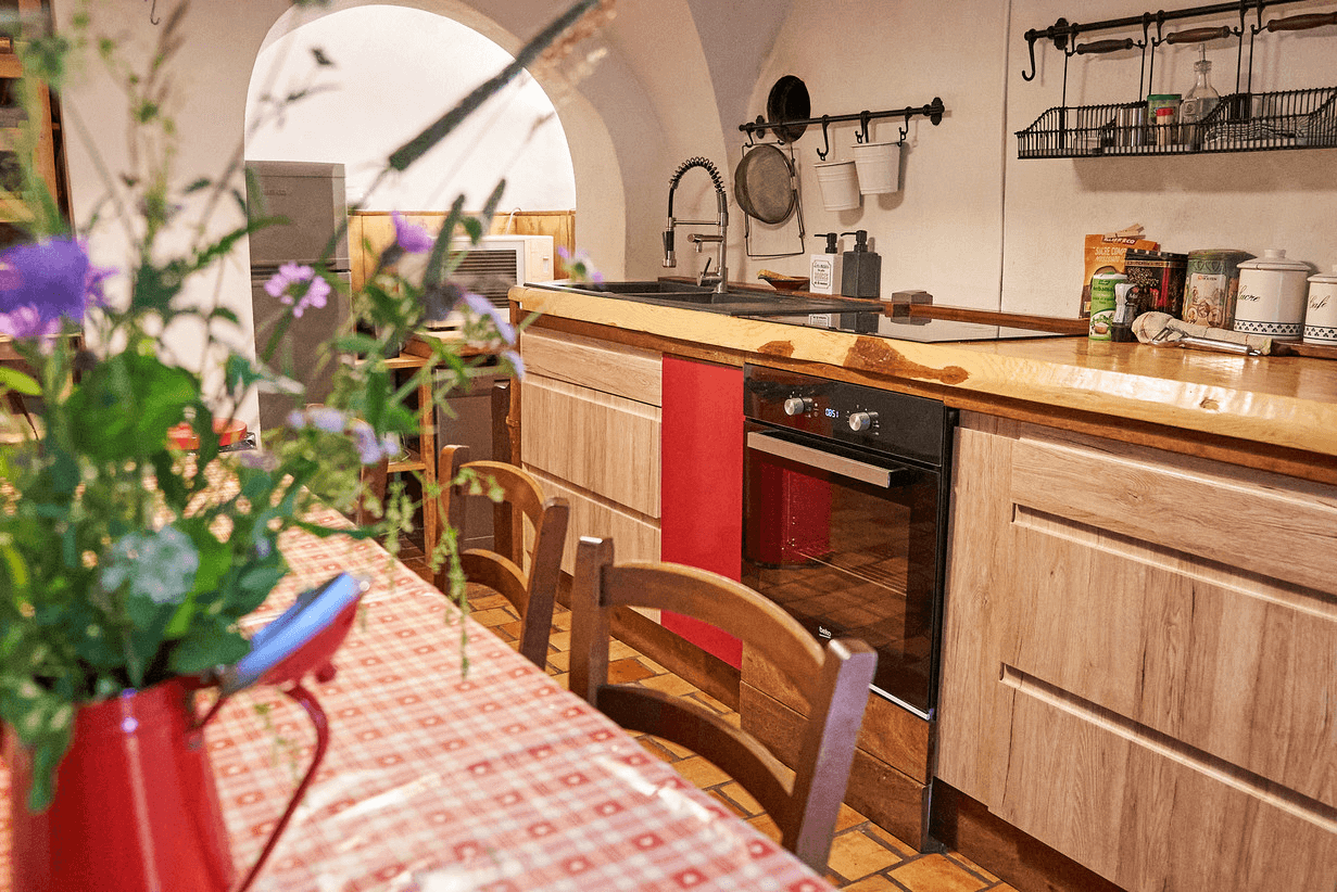 Une cuisine confortable et bien éclairée avec des armoires en bois et un accent rouge, une table à manger couverte d'une nappe à carreaux, et une plante en pot ajoutant une touche de verdure.