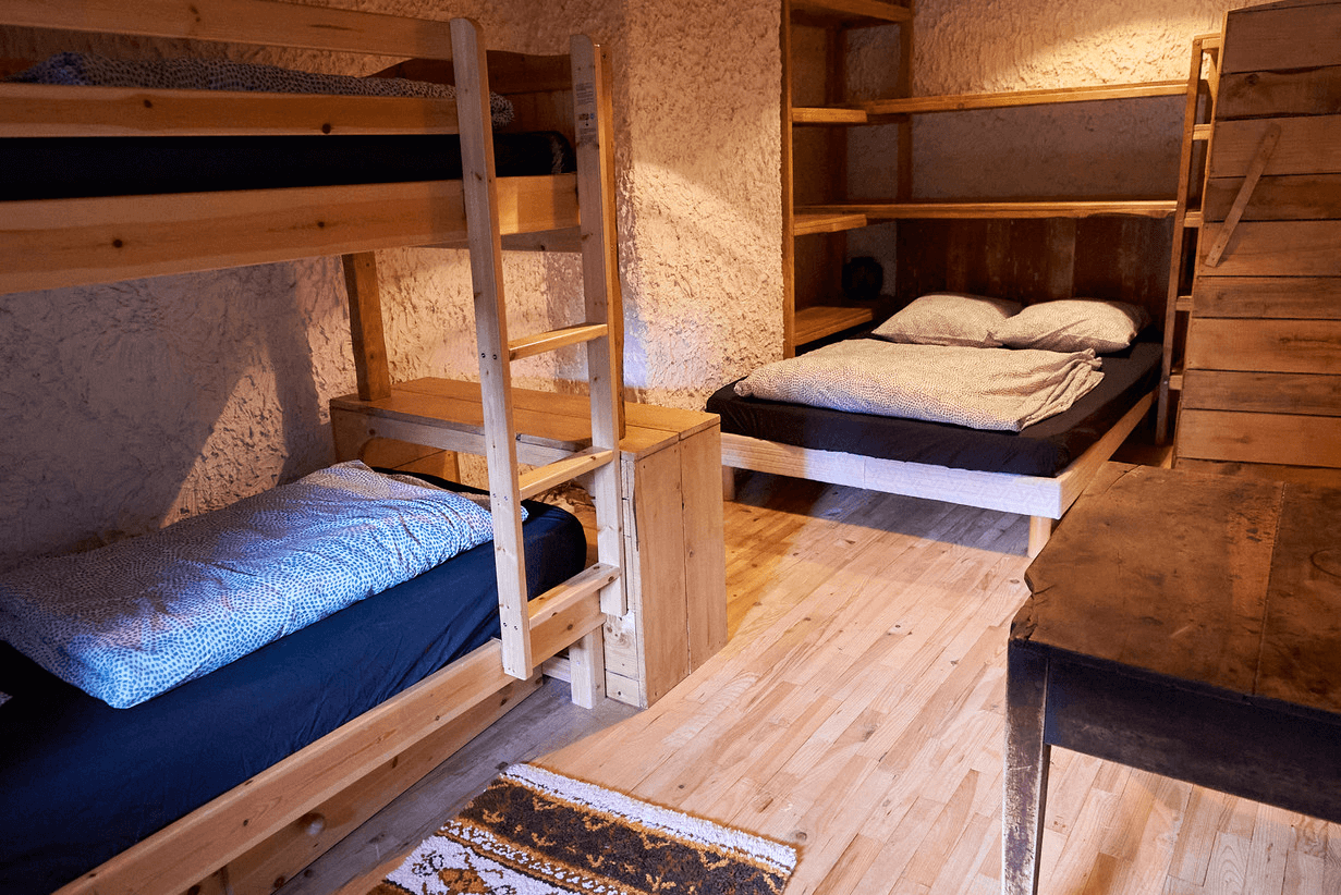 Un intérieur en bois confortable avec un lit superposé avec une literie à motifs bleus sur la gauche, un lit simple avec une literie grise sur la droite, un parquet, un petit tapis et une table en bois.