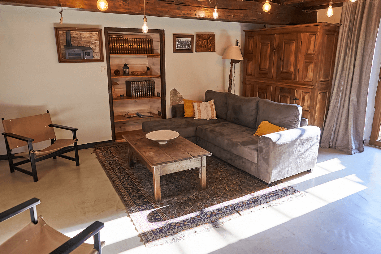 Un salon confortable avec un canapé gris orné de coussins jaunes et blancs, une table basse en bois sur un tapis orné, des chaises en bois, et un éclairage d'ambiance qui projette une lueur chaleureuse, mettant en valeur l'esthétique rustique de la pièce.