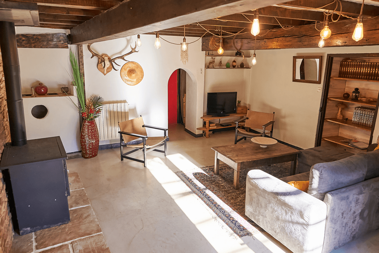 Un salon confortable et bien éclairé présentant un mélange d'éléments modernes et rustiques, avec des poutres en bois au plafond, une variété d'options de sièges comprenant des canapés et des chaises, des objets décoratifs sur les murs, et une télévision.