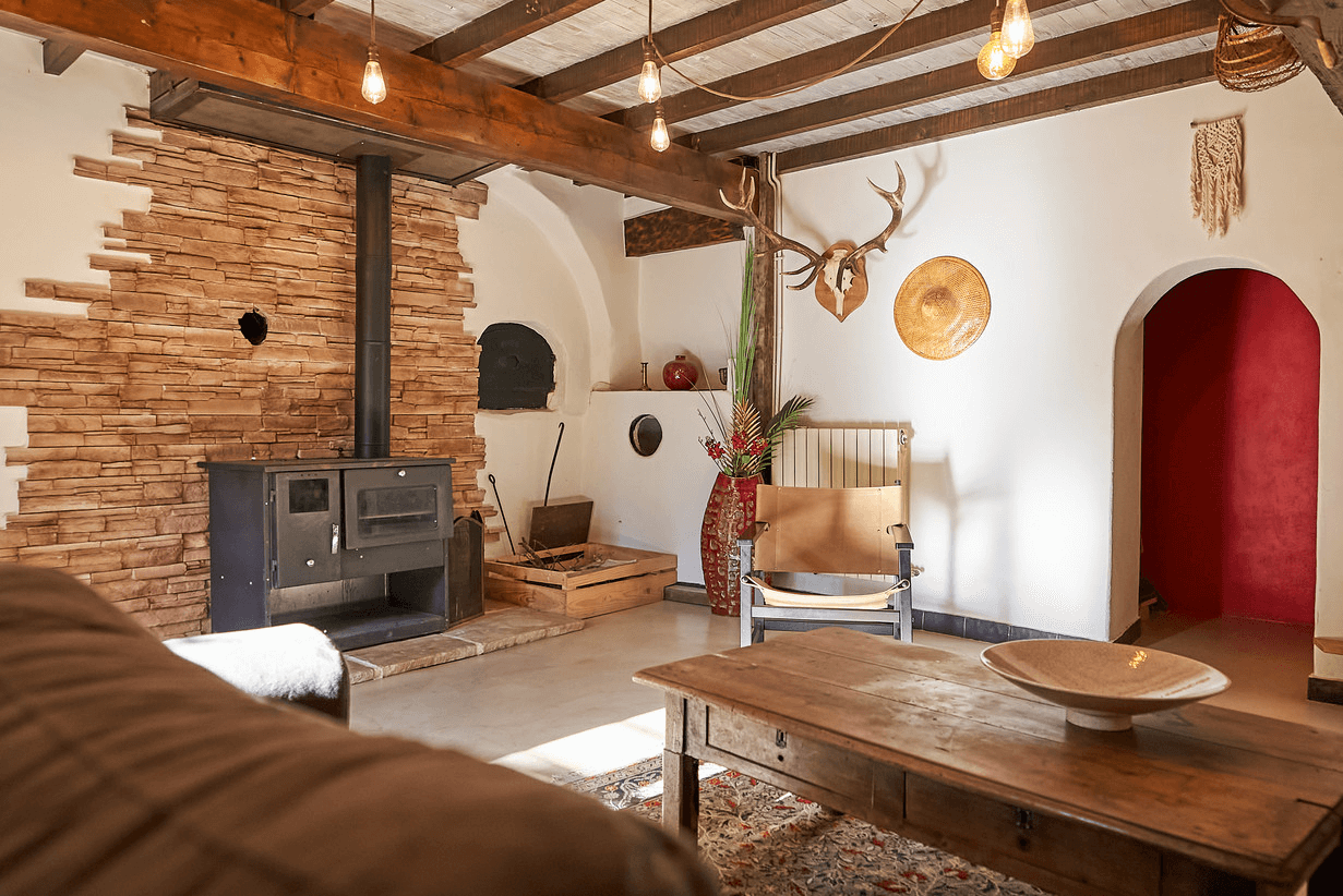Un salon rustique et confortable avec un poêle à bois contre un mur en pierre empilée, des poutres en bois au plafond, une table et une chaise en bois vintage, des objets décoratifs comprenant des cornes et des paniers tissés accrochés aux murs, et une porte rouge voûtée menant à une autre pièce.