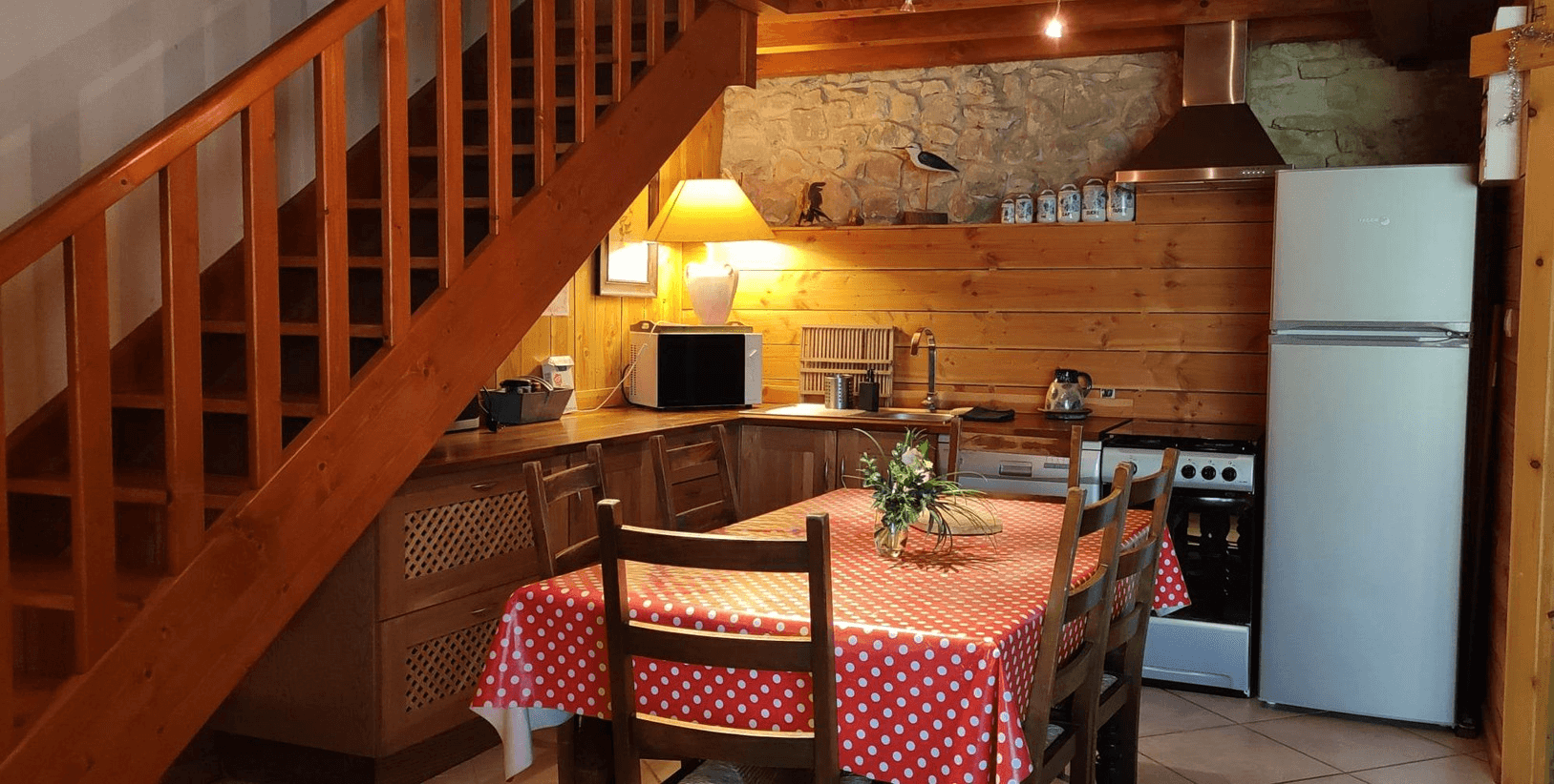Une cuisine confortable et bien éclairée avec un thème en bois, comprenant une table à manger recouverte d'une nappe rouge à pois blanc, entourée de chaises, adjacente à un escalier en bois menant à l'étage; la cuisine est équipée d'appareils modernes et a des accents de mur en pierre.
