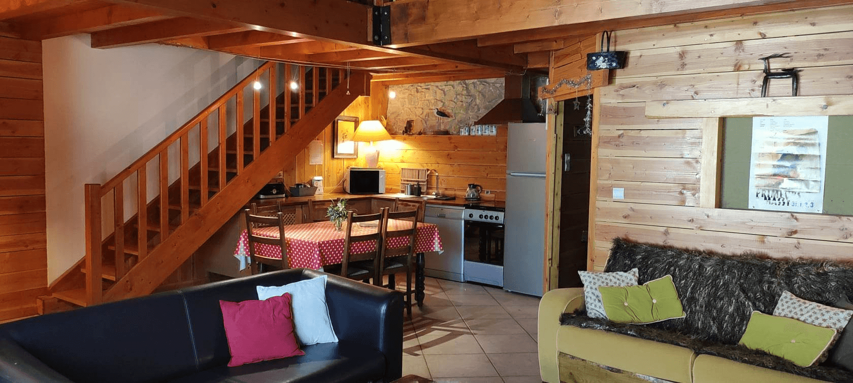 Un intérieur confortable et bien éclairé d'une cabane en bois comprenant un salon avec un canapé foncé orné de coussins colorés, adjacent à une salle à manger avec une nappe à carreaux rouge et blanc, le tout sous la lueur chaleureuse des lumières du plafond; un escalier en bois mène à un niveau supérieur.