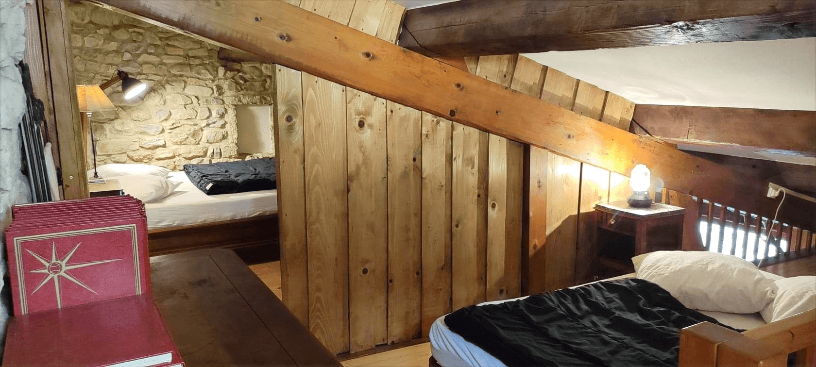 Une chambre rustique et confortable avec deux lits aux draps blancs et noirs, des poutres en bois apparentes, des murs en pierre, et deux lampes allumées créant une ambiance chaleureuse.