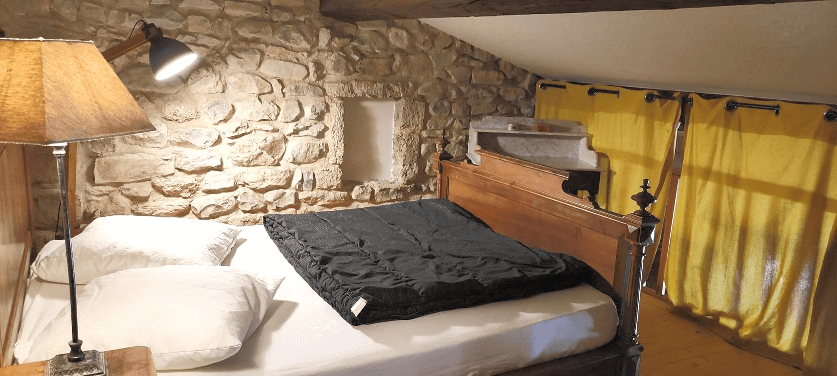 Une chambre confortable avec un lit aux draps blancs et une couverture foncée, placé contre un mur en pierre rustique, éclairé par une lampe sur pied; des rideaux jaunes couvrent les fenêtres à droite.