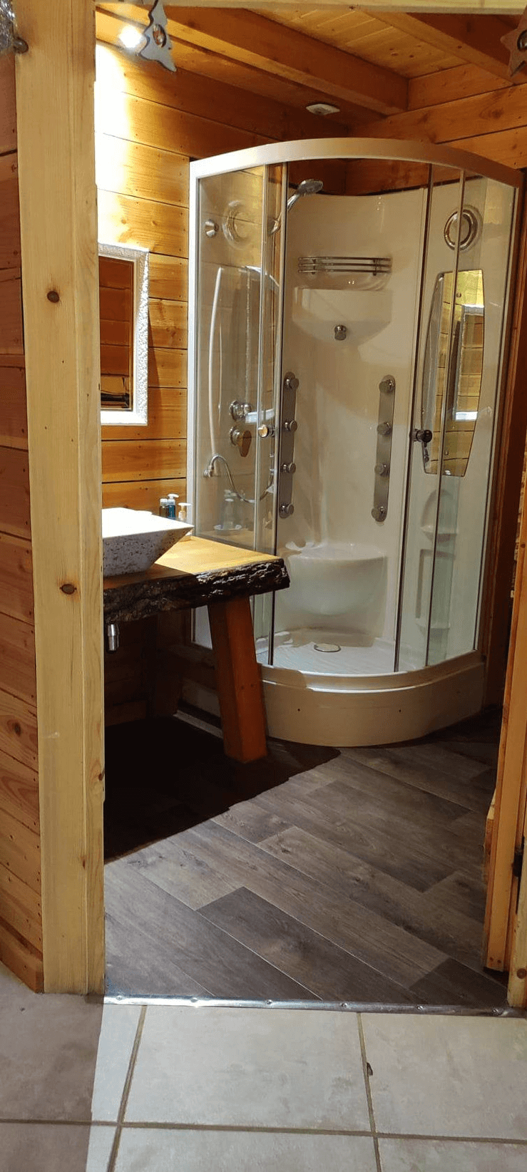 Une salle de bain confortable avec une cabine de douche moderne en verre courbé, un tabouret en bois avec une serviette, et des murs en bois qui lui donnent une atmosphère chaleureuse et accueillante.