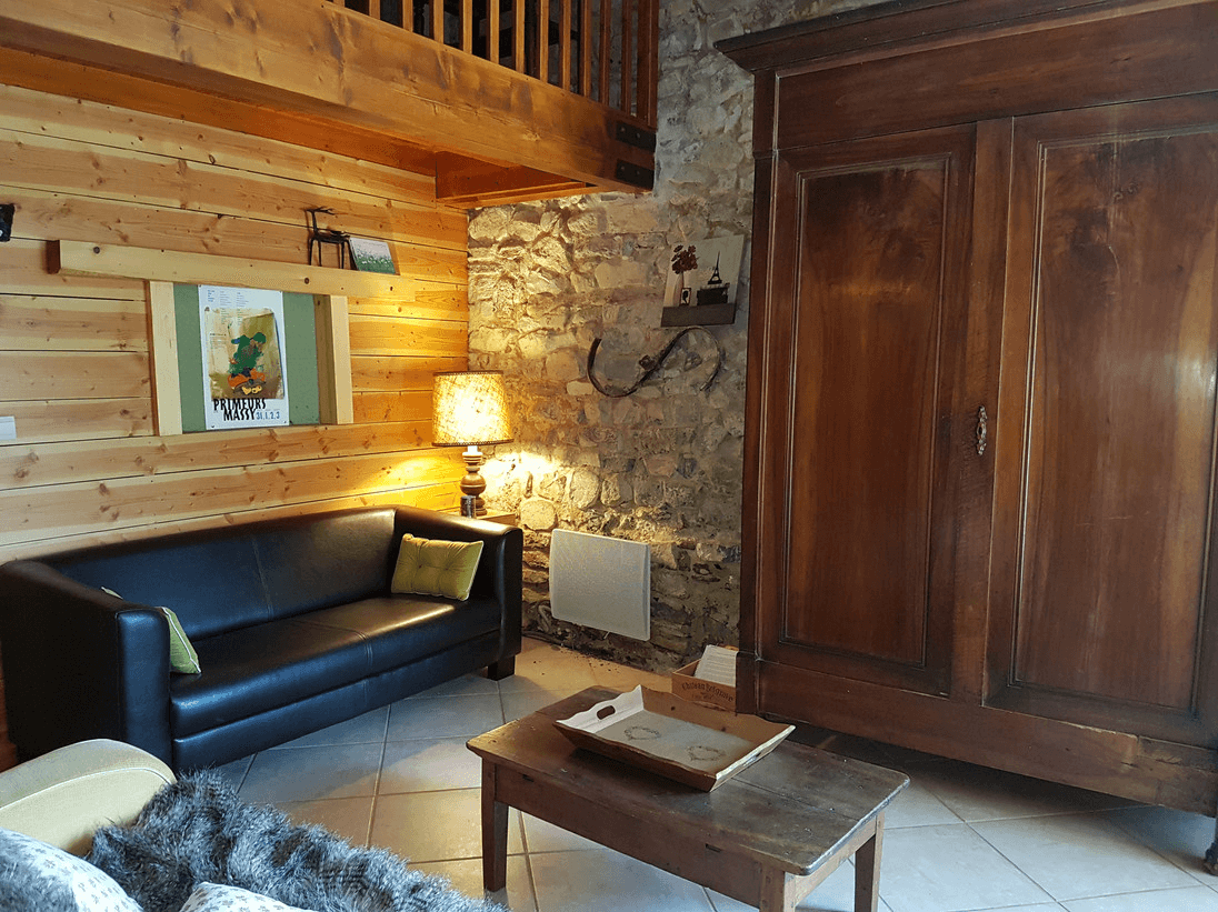 Un intérieur confortable avec un mélange d'éléments modernes et rustiques, comprenant un canapé en cuir noir, des murs et des meubles en bois, un mur en pierre, des œuvres d'art sur le mur, et un éclairage chaleureux provenant d'une lampe de table.