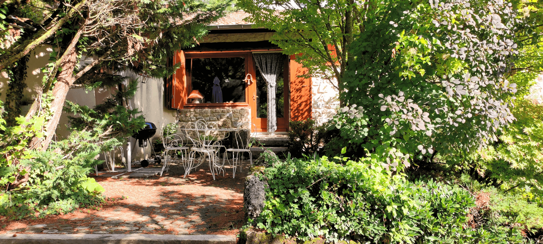Une maison entourée de verdure luxuriante avec un patio comprenant des meubles de jardin, éclairée par la lumière du soleil.
