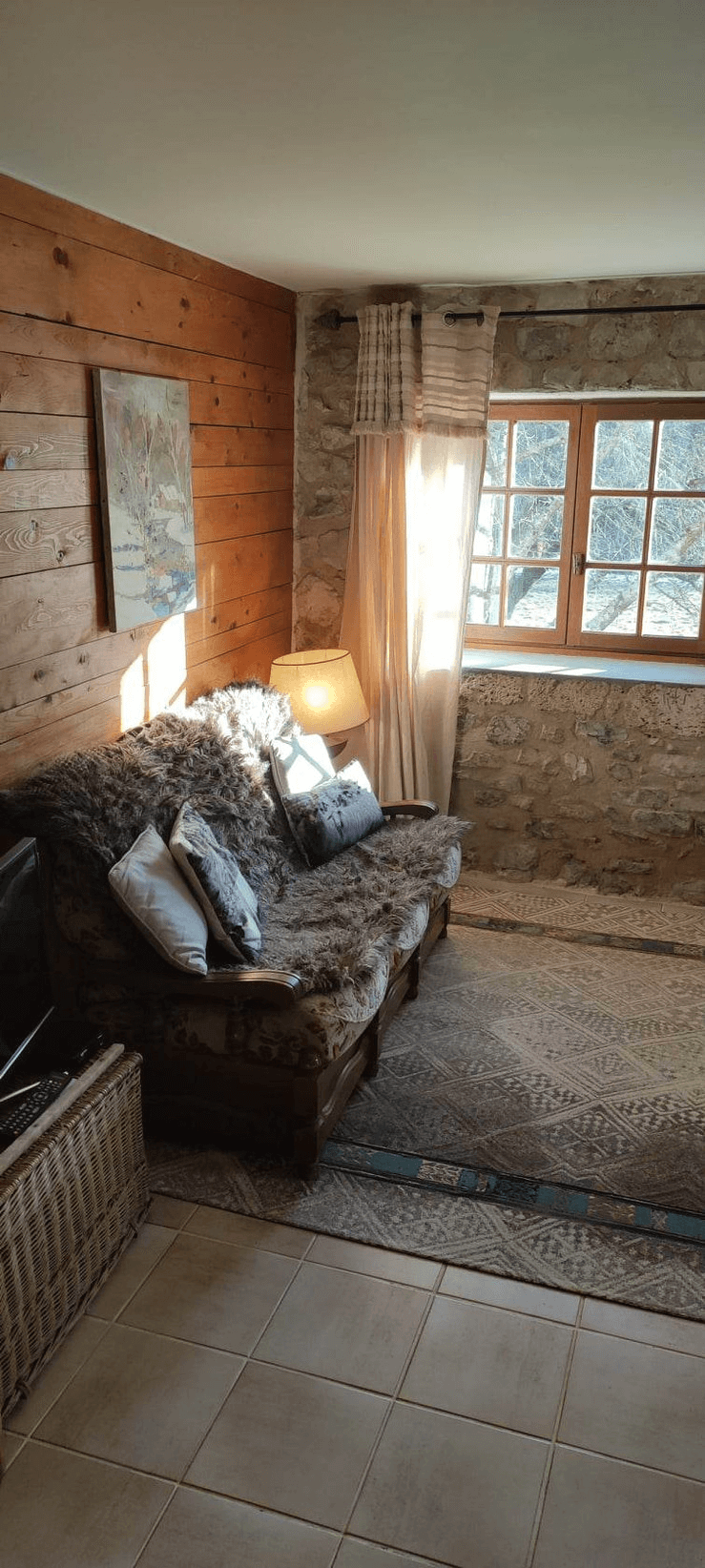 Une pièce confortable avec un canapé recouvert de coussins et une couverture, une lampe allumée à côté, un tapis sur le sol carrelé, des murs en bois et en pierre, et une fenêtre laissant entrer la lumière du soleil.