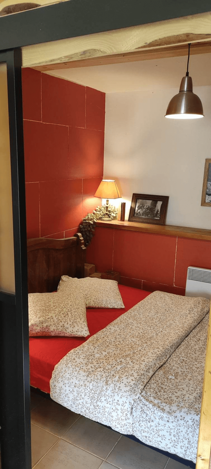 Une chambre confortable avec un lit double couvert d'une literie à motifs floraux, des murs rouges et blancs, une lampe de chevet allumée et un luminaire suspendu éclairant la pièce.