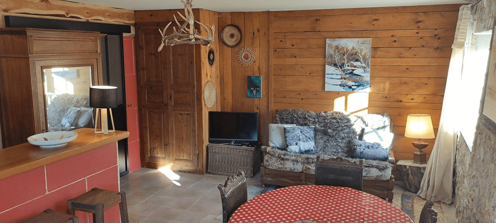 Une pièce chaleureuse avec des murs en bois, un canapé recouvert d'une couverture en fausse fourrure, une table ronde avec une nappe rouge à pois blancs, et divers éléments décoratifs comme un tableau, des lampes et des objets sur les murs.