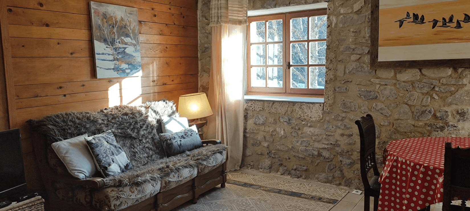 Une pièce confortable avec un canapé recouvert de coussins et une couverture, une table à côté avec une lampe allumée, des œuvres d'art aux murs, une fenêtre laissant entrer la lumière naturelle et une table à manger avec des chaises.