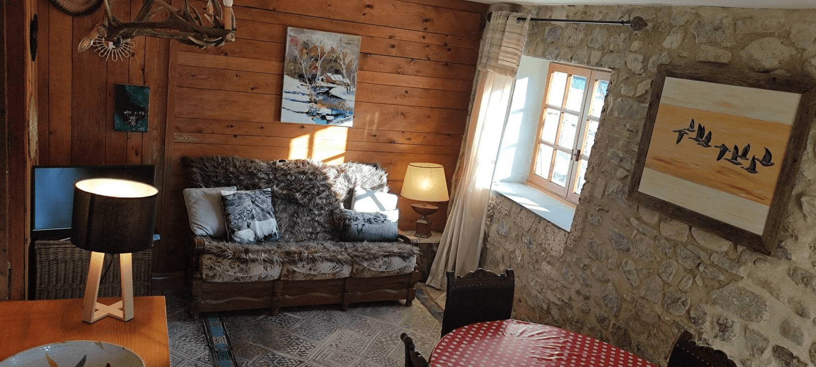 Une pièce confortable avec un canapé recouvert de coussins et une couverture, entourée d'œuvres d'art accrochées aux murs en bois et en pierre, éclairée par la lumière naturelle provenant d'une fenêtre et des lampes allumées.