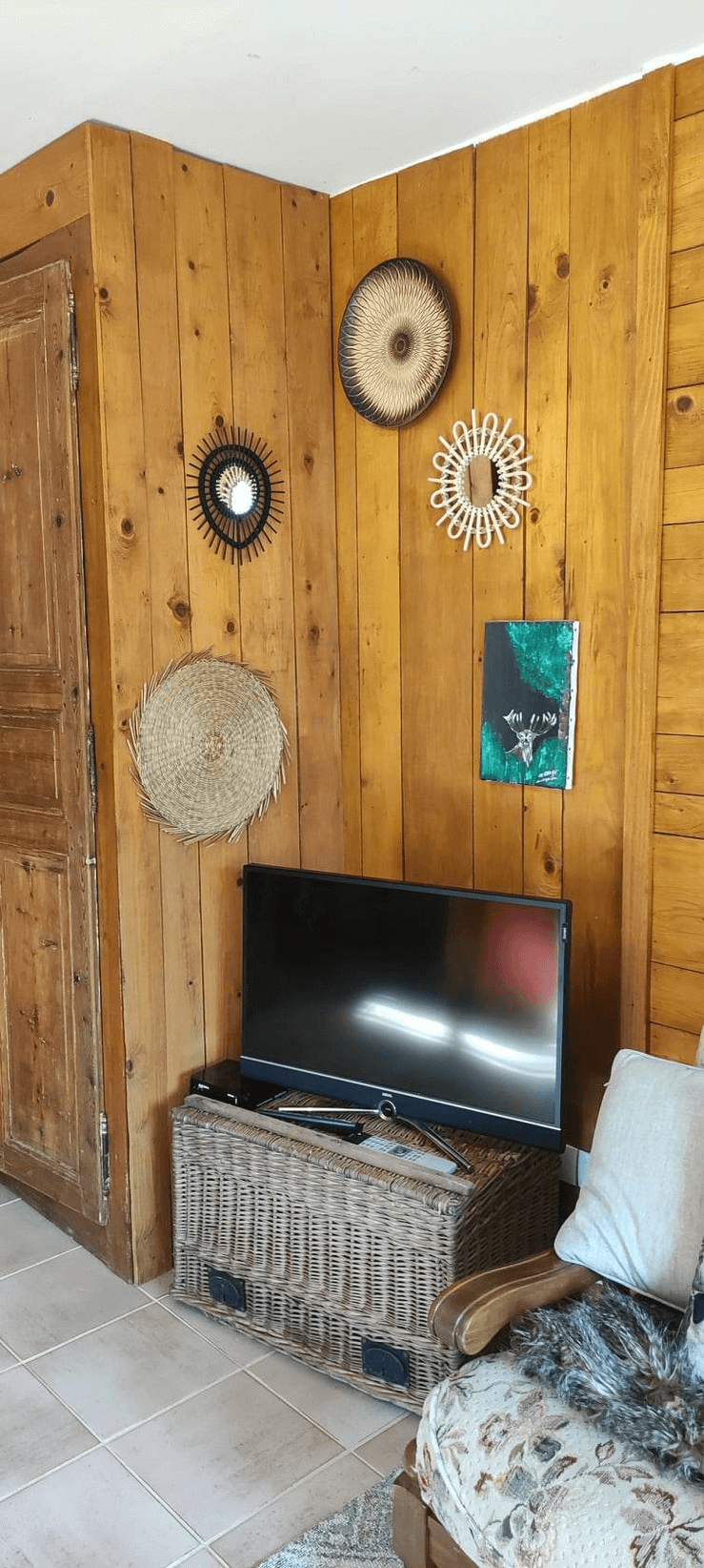 Une pièce avec un mur en bois sur lequel sont accrochés trois décorations murales circulaires et une petite peinture, avec une télévision noire posée sur un panier en osier à côté d'un canapé.